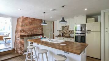 A modern kitchen with a brick fireplace, wooden island, white bar stools, and stainless steel appliances.