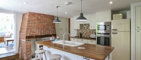 A modern kitchen with a brick fireplace, wooden island, white bar stools, and stainless steel appliances.