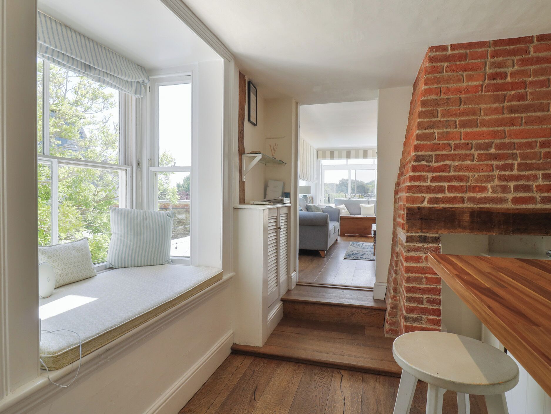 A room with a built-in bench by a large window, a brick fireplace, and a view into another room with a sofa.