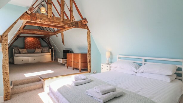 Un dormitorio con techo inclinado, una cama, un aparador y un pequeño rincón de descanso con pared de ladrillo.