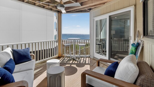 Een balkon met uitzicht op zee, een witte bank met blauwe kussens, een klein rond tafeltje en een rieten stoel.