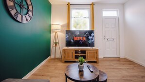 Een woonkamer met een grote flatscreen televisie, een ronde houten salontafel, een potplant, een vloerlamp en een wandklok.