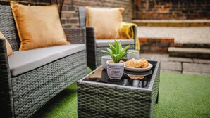 Wicker meubels met kussens, een tafeltje met een plant en croissant, en een bakstenen muur op de achtergrond.