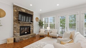 A living room with a stone fireplace, white furniture, and large windows with blinds. - Lakefront Retreat • Private Dock + Firepit + Game Room | Montclair Manor (Mooresville)
