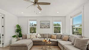 A modern living room with a sectional sofa, a small round table with a bottle, and a ceiling fan. - Sleeps 32 • Private Pool + Game Room + Golf Cart | Grayton Dreams (Santa Rosa Beach)