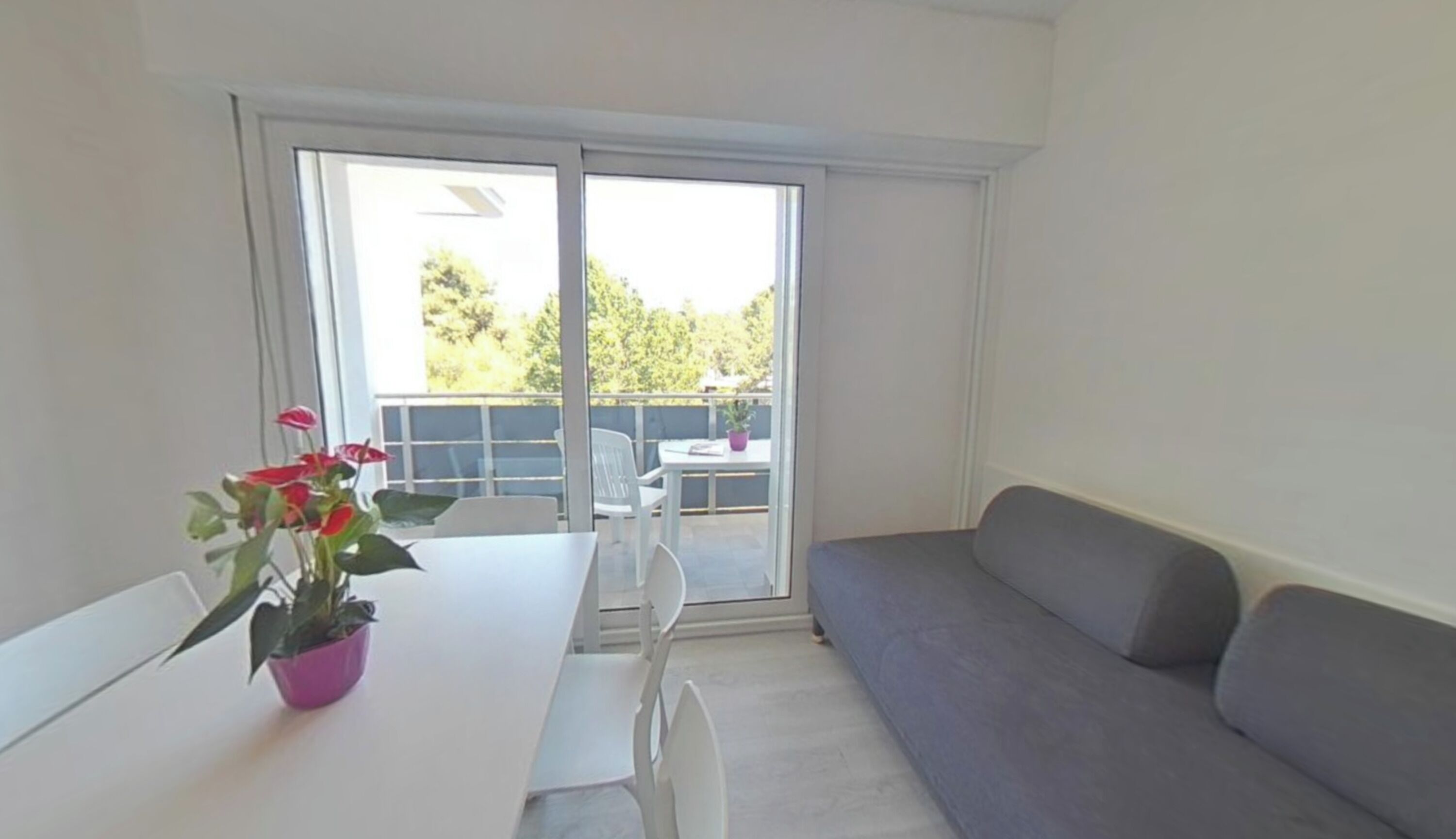 Ein Zimmer mit einem weißen Tisch, einem grauen Sofa und einer Glastür, die zu einem Außenbereich mit Tisch und Stühlen führt.