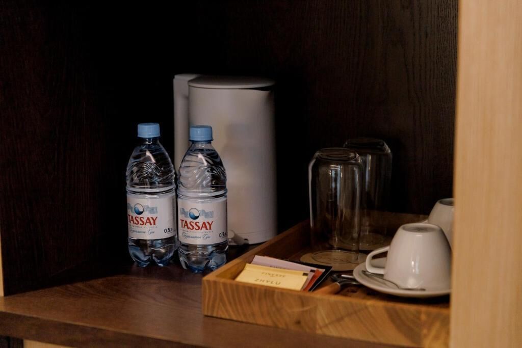 A hotel room with a minibar containing two bottles of TASSAY water, a coffee maker, and a tray with white cups and a card.