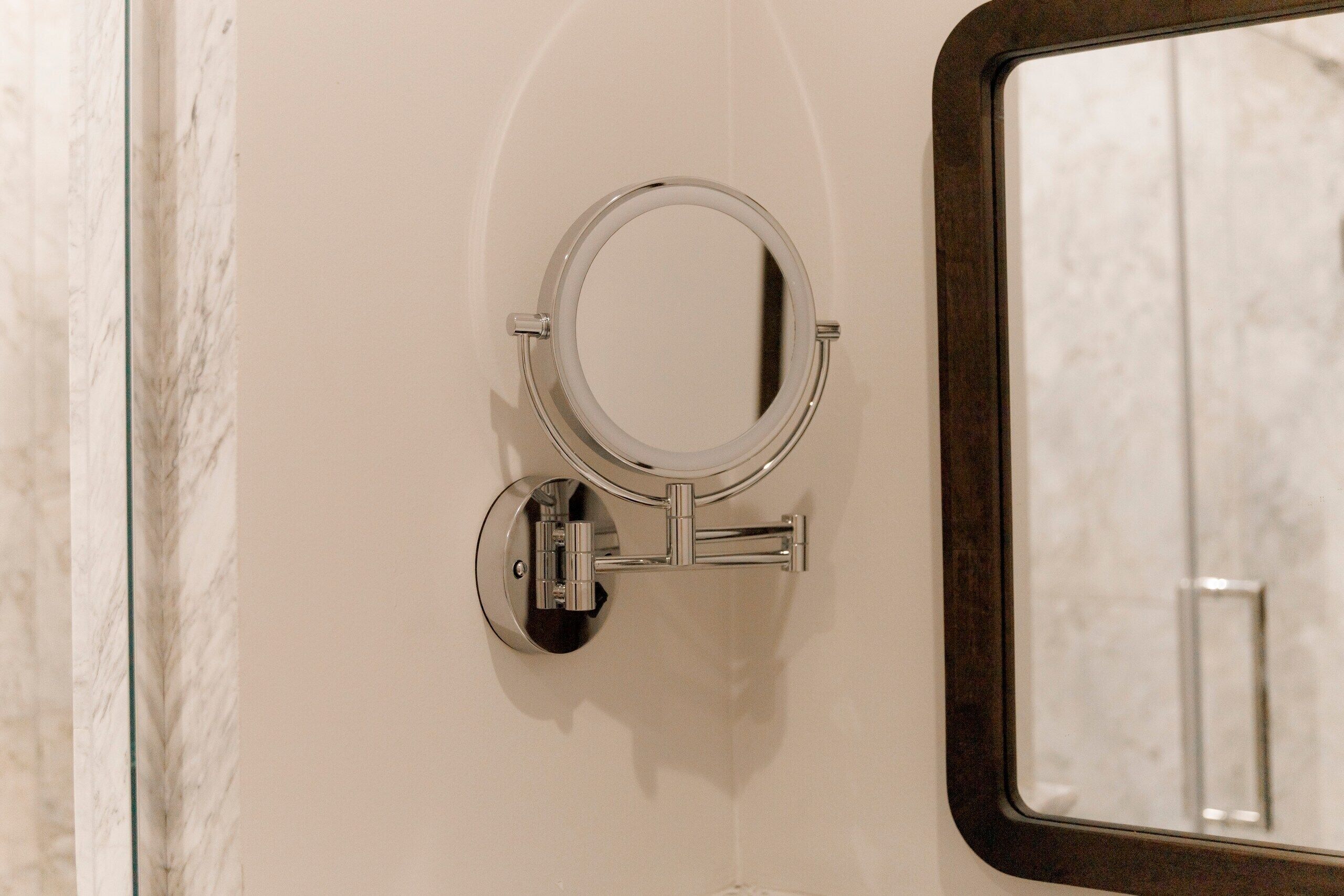 Un espejo instalado en la pared, con un marco cromado, un objeto rectangular pequeño debajo y el reflejo del interior de un baño.