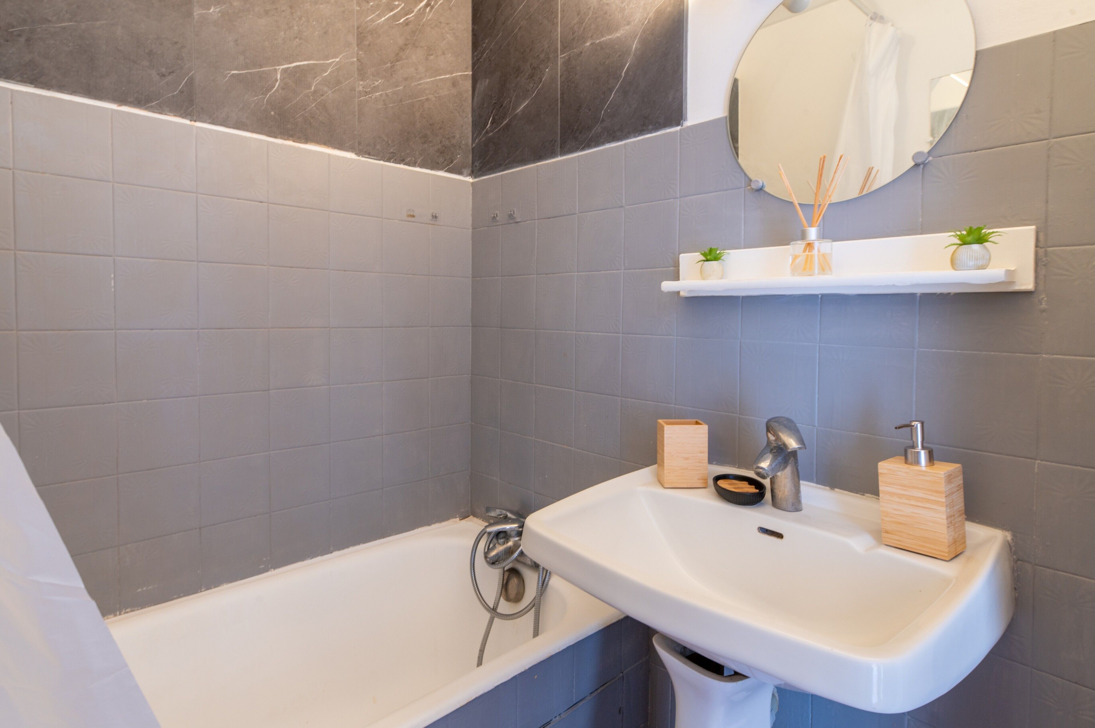 A bathroom with a bathtub, a sink, a mirror, and a shelf with a plant and a soap dispenser.
