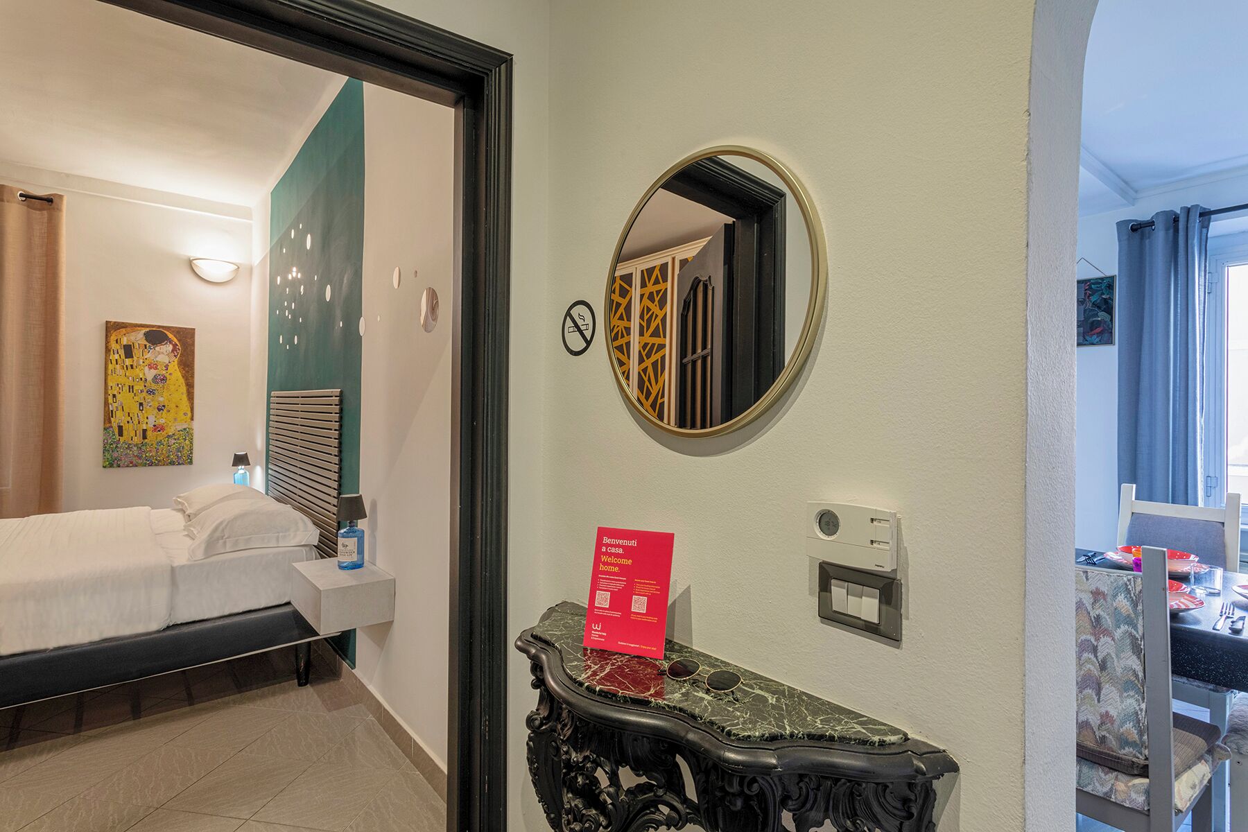 A hotel room with a bed, a bedside table with a lamp, a mirror, and a sign that says 'Welcome home.'