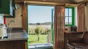 A room with a kitchen counter, a dining area, a leather chair, and a view of a green field through an open door. - Luxury Shepherd Hut - The White Princess (Bolton Percy)