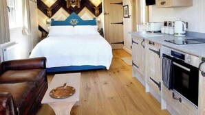 A compact room with a bed, a wooden table, a kitchenette, and a ceiling light fixture. - Luxury Shepherd Hut - The White Princess (Bolton Percy)