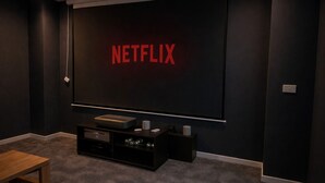 Tummasävyinen huone, jonka seinälle on asennettu suuri, litteänäyttöinen televisio, jossa näkyy punaisilla kirjaimilla teksti ”NETFLIX”.