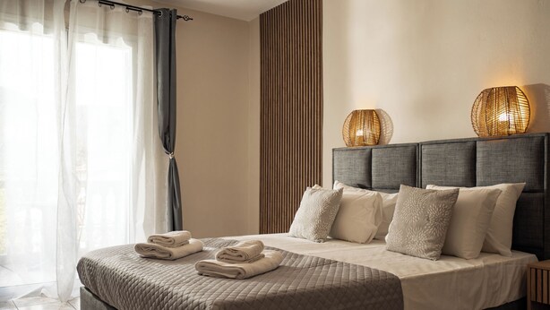 A neatly made bed with pillows and folded towels, a bedside lamp, and a view of the outdoors through a window with curtains. - Aggire Summer Suites (Zakynthos)