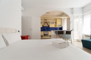 Una camera d'albergo con un letto, una poltrona rossa, un angolo cottura visibile tramite un arco e una finestra con tende.