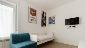 Een kamer met een blauwgroene bank, een wit bed, een televisie en twee posters aan de muur.