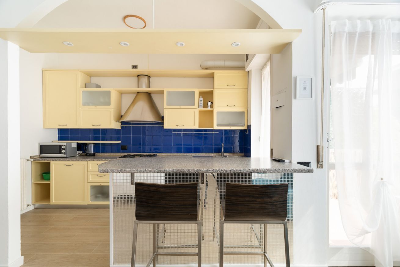 A compact kitchen with yellow cabinets, a blue backsplash, and a small island with two bar stools.