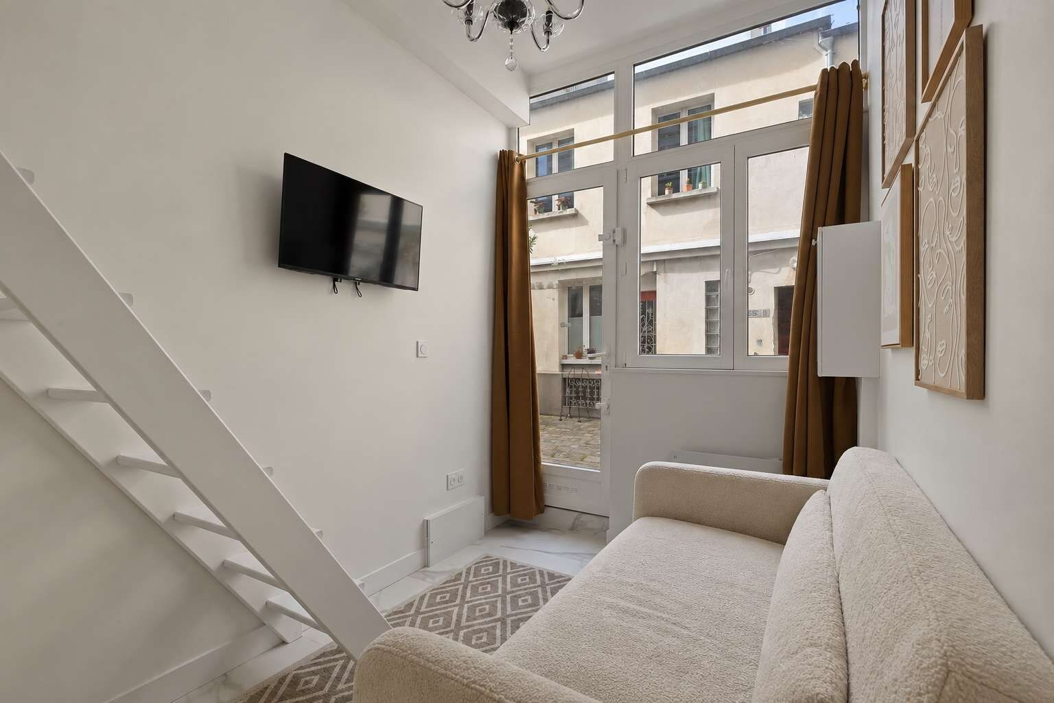 Sala de estar moderna com televisão de tela plana, sofá, escada e vista para uma varanda externa através de uma porta de vidro.