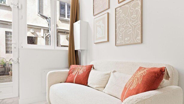 Un canapé blanc agrémenté de coussins rouges et beige, une fenêtre avec des rideaux et des œuvres d’art encadrées accrochées au mur.