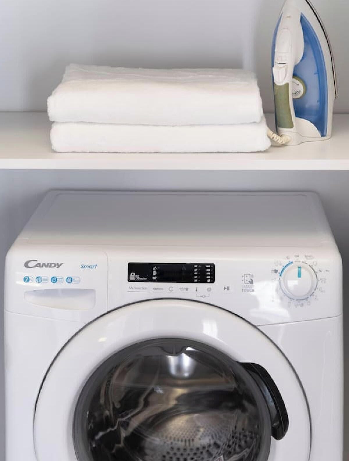 A white washing machine with a digital display and control panel, a stacked white towel on a shelf, and a blue and white iron on the same shelf.