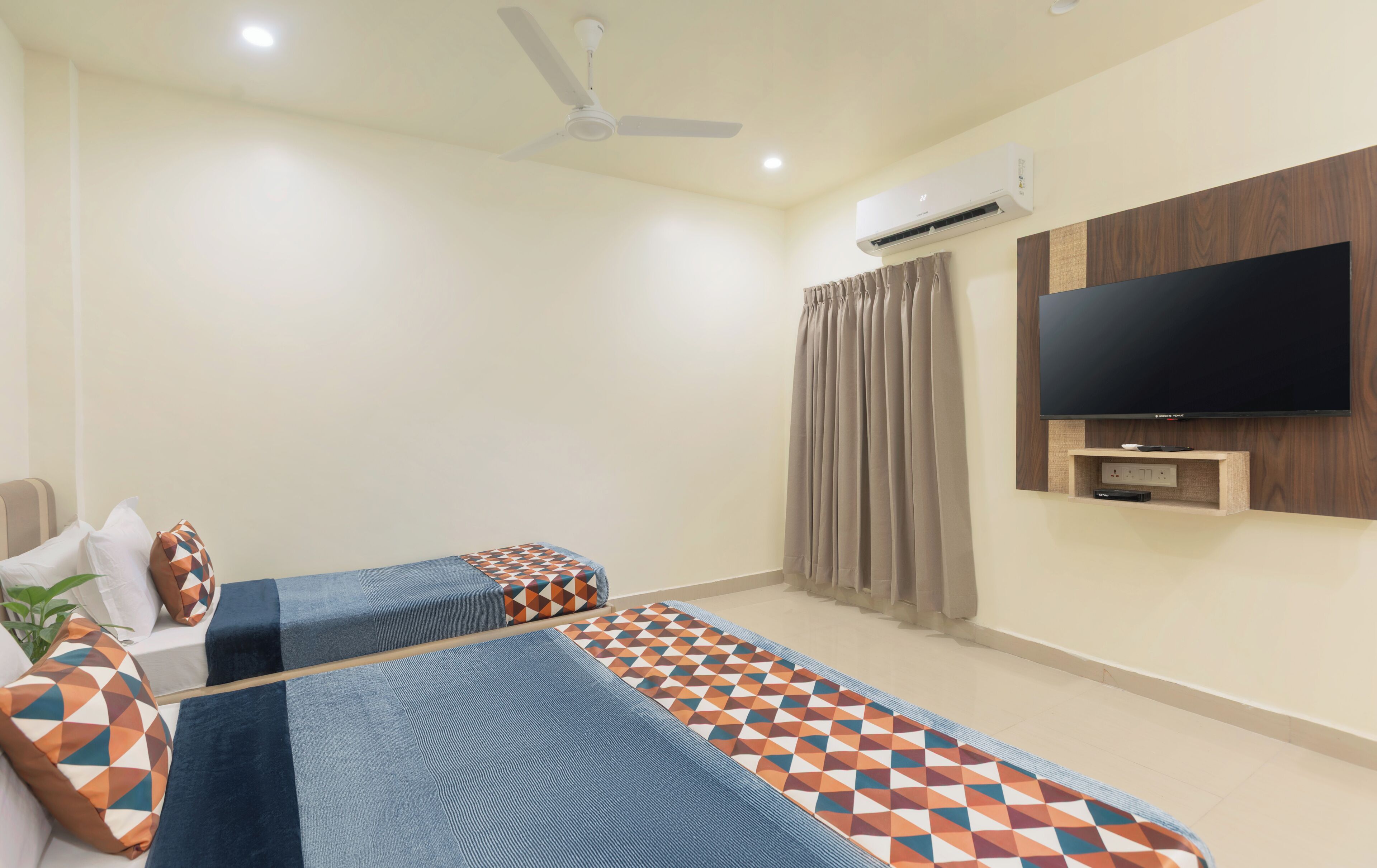 Habitación de hotel con dos camas, ventilador de techo, unidad de aire acondicionado, televisor montado en la pared y ventana con cortinas.
