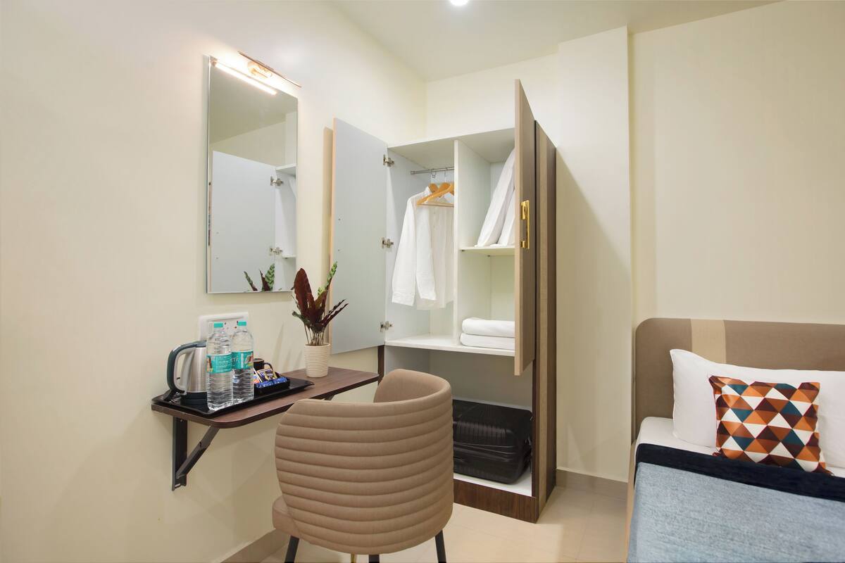 A hotel room with a bed, a desk with a chair, a wardrobe, a mirror, and a small table with toiletries.