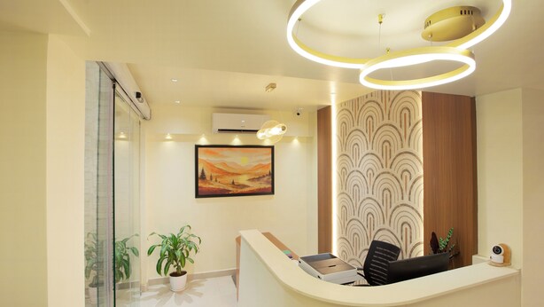A reception area with a curved desk, a potted plant, a painting, and a modern ceiling light fixture. - Greens Venue (Chennai)