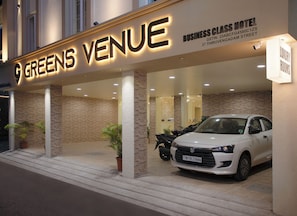 Entrada de un hotel con un cartel que dice 'Greens Venue' y 'Business Class Hotel'. Hay plantas en macetas y un coche blanco estacionado afuera.