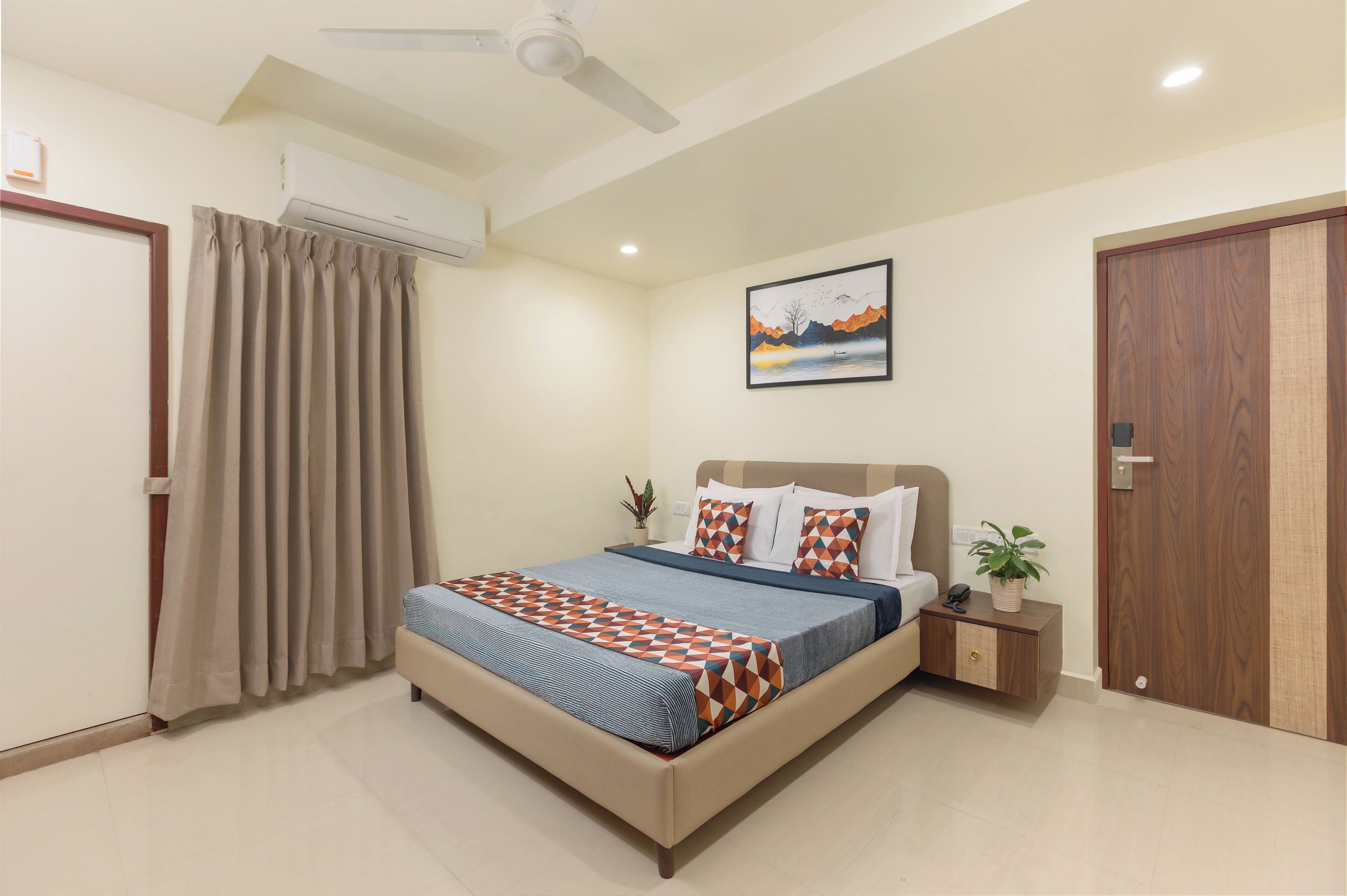 A bedroom with a bed, curtains, a ceiling fan, an air conditioning unit, a nightstand with a plant, and a wooden door.