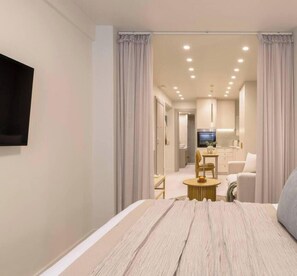 A modern hotel room with a large bed, a TV mounted on the wall, a dining area with a table and chairs, and a kitchen in the background. - Chic Stay | Spa-Style Bath | Acropolis (Athens)