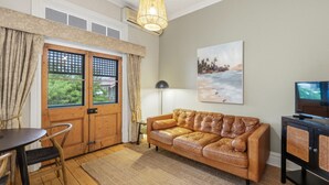A living room with a leather sofa, a dining table with chairs, a TV stand, and a large window with curtains. - Apartment Faro - Heart of South Freo with Parking (South Fremantle)