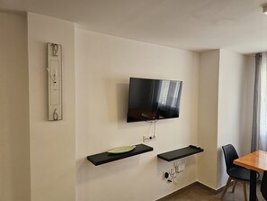 Un salon moderne avec un téléviseur à écran plat fixé au mur, deux étagères noires murales, une table à manger en bois avec des chaises, et un petit objet décoratif placé à gauche.