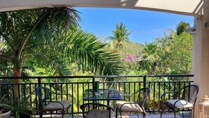 Une terrasse dotée d’une table et de chaises, entourée d’une balustrade, et offrant une vue sur un jardin luxuriant où poussent différentes plantes et arbres.