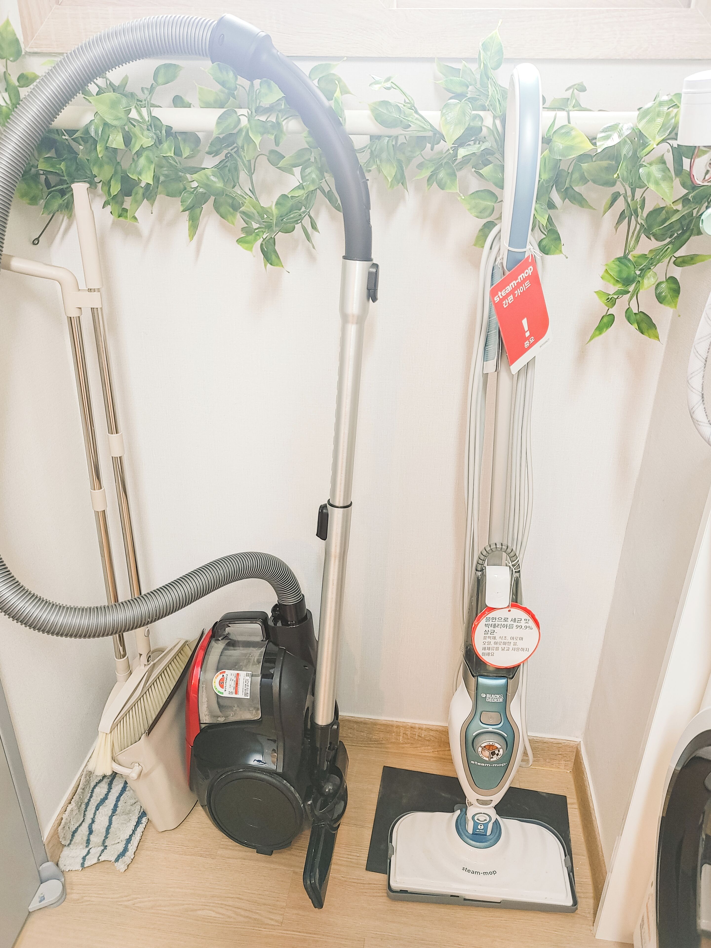 Un aspirateur, une serpillière et un balai sont posés dans un coin, devant un plancher en bois et un mur blanc.