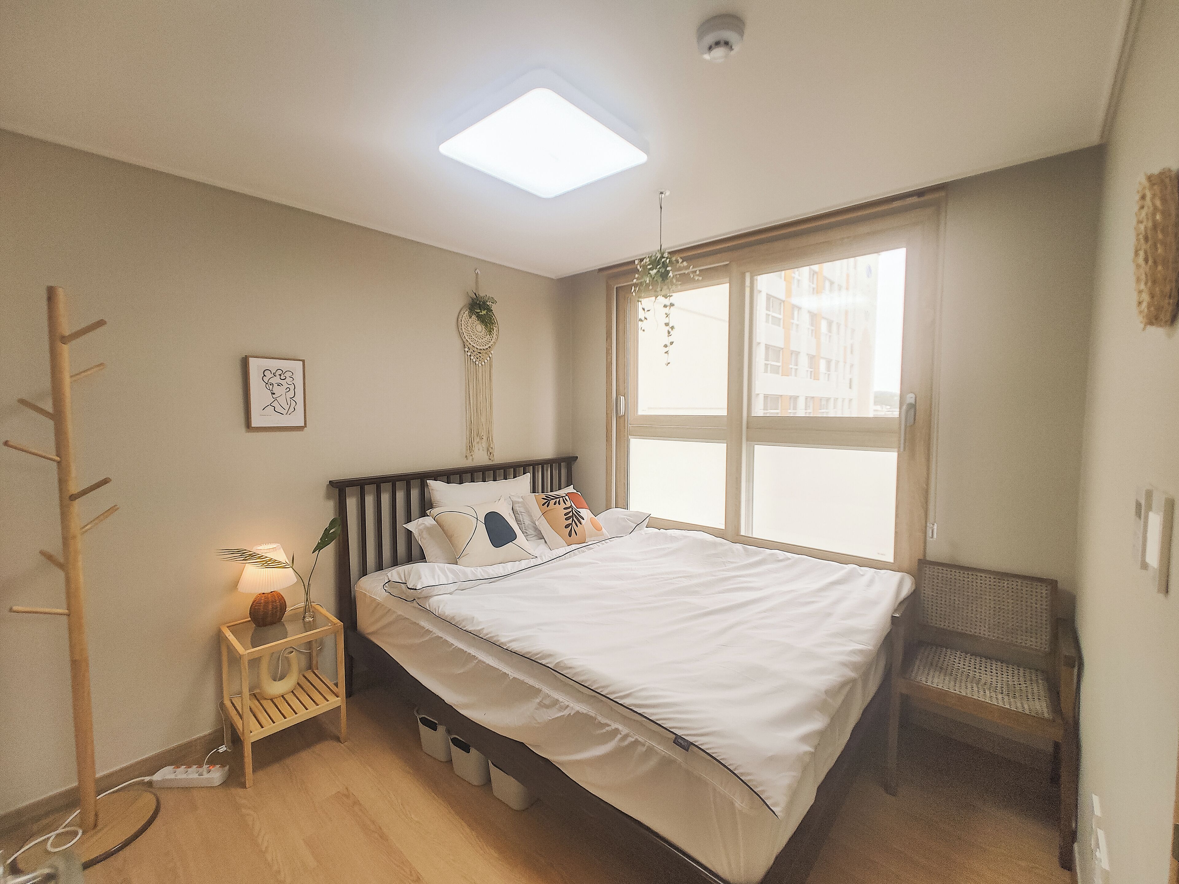 Un dormitorio con cama, mesita de noche, lámpara, ventana y vistas a edificios.