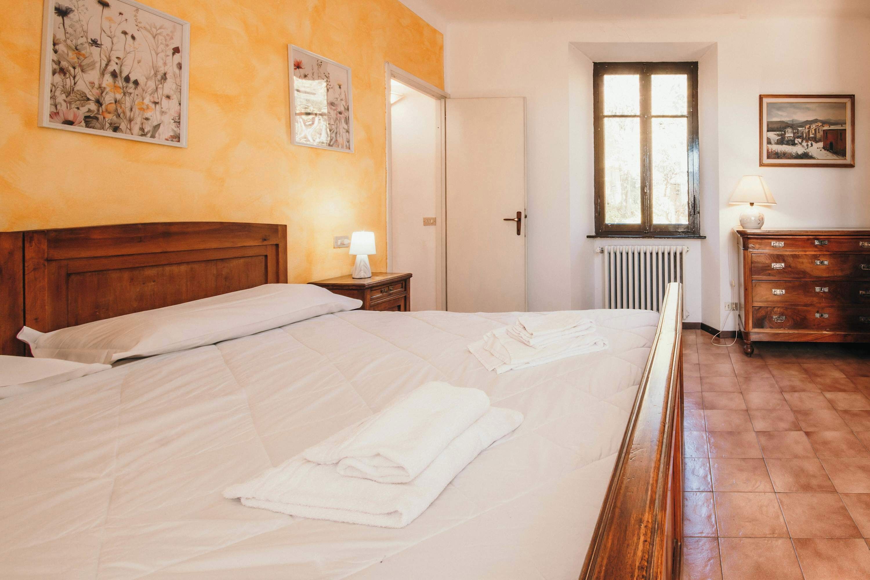 Ein Hotelzimmer mit Bett, Nachttisch, Kommode, Heizkörper und zwei gerahmten Bildern an der Wand.