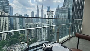 Un balcon avec une table et une chaise en verre, donnant sur un paysage urbain composé d’immeubles de grande hauteur et d’un parking.