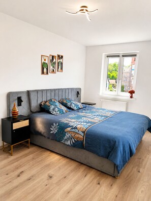 Ein modernes Schlafzimmer mit einem grau gepolsterten Bett, einem Nachttisch mit einer Vase und einem Fenster mit Blick auf Grünflächen.