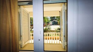 Een kamer met twee openslaande deuren die uitkomen op een balkon met een witte reling en uitzicht op een huis en groen.