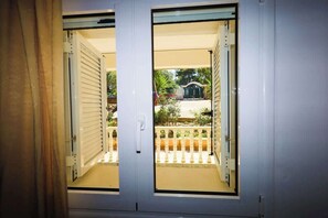 A room with two open sliding doors leading to a balcony with a white railing and a view of a house and greenery. - Charming Country House, Just Steps from Everything (Kiparissia)