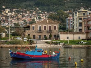 Un bâtiment en pierre avec un toit en tuiles, un bateau amarré à un quai et une colline parsemée de bâtiments résidentiels.