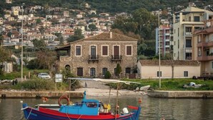 A stone building with a tiled roof, a boat docked at a pier, and a hillside with residential buildings. - Kyparissia Aries Stone Maisonette, Panoramic Views (Kiparissia)