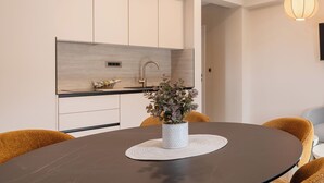 Moderni keittiö, jossa on valkoiset kaapit, harmaa työtaso ja pesuallas. Keittiössä on pyöreä ruokapöytä, jonka päällä on valkoinen alusta ja pöydällä on ruukkukasvi.