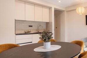 A modern kitchen with white cabinets, a grey countertop, and a sink. There's a round dining table with a white placemat and a potted plant. - Villa Merkur-apartment 3 (Makarska)