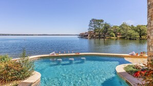 Una piscina con agua azul cristalina, rodeada de un muro de piedra y plantas, con vista al mar tranquilo y a una costa lejana con árboles y edificios.