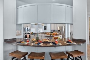 A modern kitchen with a curved island, bar stools, and a central island with food and drinks. - Bay Point Oasis  | Concierge Services (Horseshoe Bay)