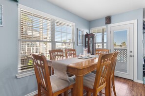 A dining room with a wooden table, six chairs, a chandelier, and a view through the window blinds.