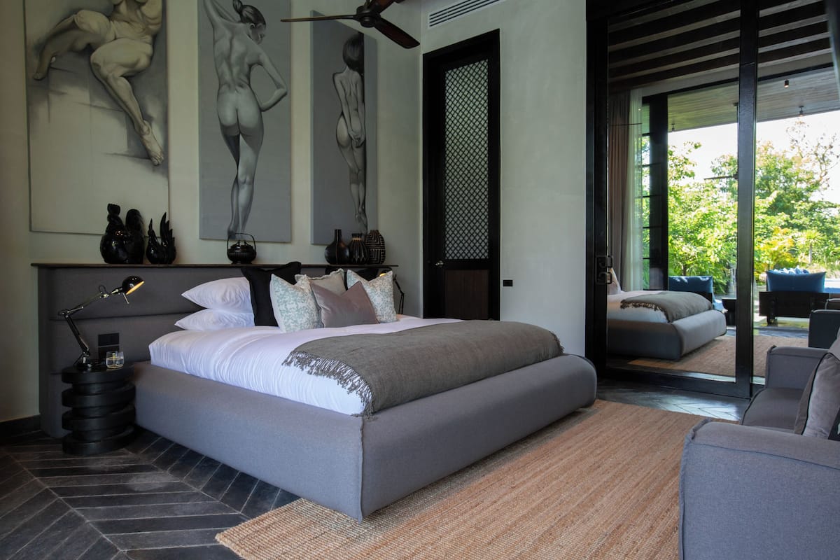 Quarto moderno com uma cama grande, uma mesa de cabeceira, uma escrivaninha e uma cadeira. Há quadros abstratos na parede e vista para o exterior através de uma porta de vidro.