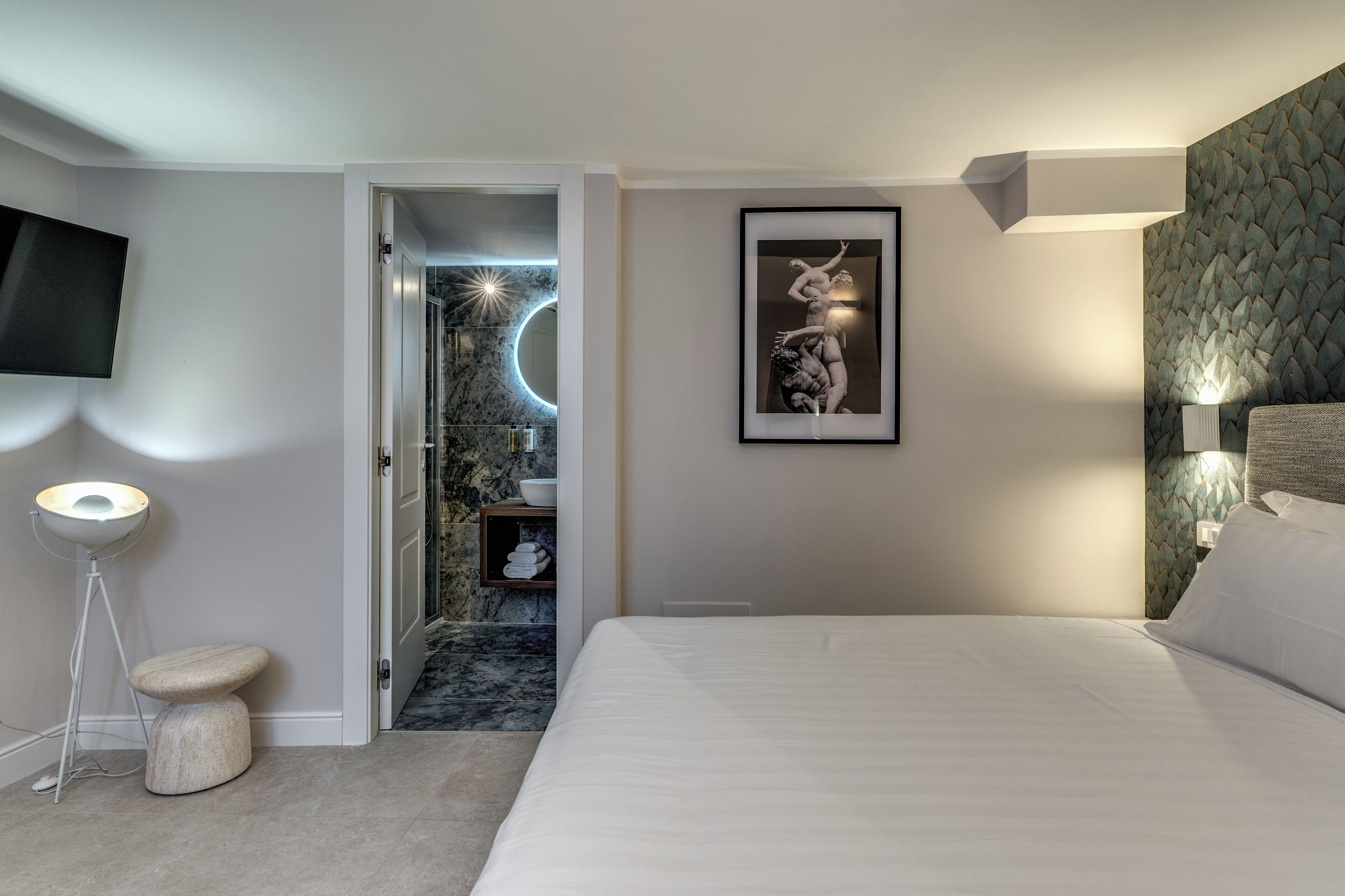 A modern hotel room with a bed, a flat-screen TV, a framed artwork, a floor lamp, and a marble bathroom visible through an open door.
