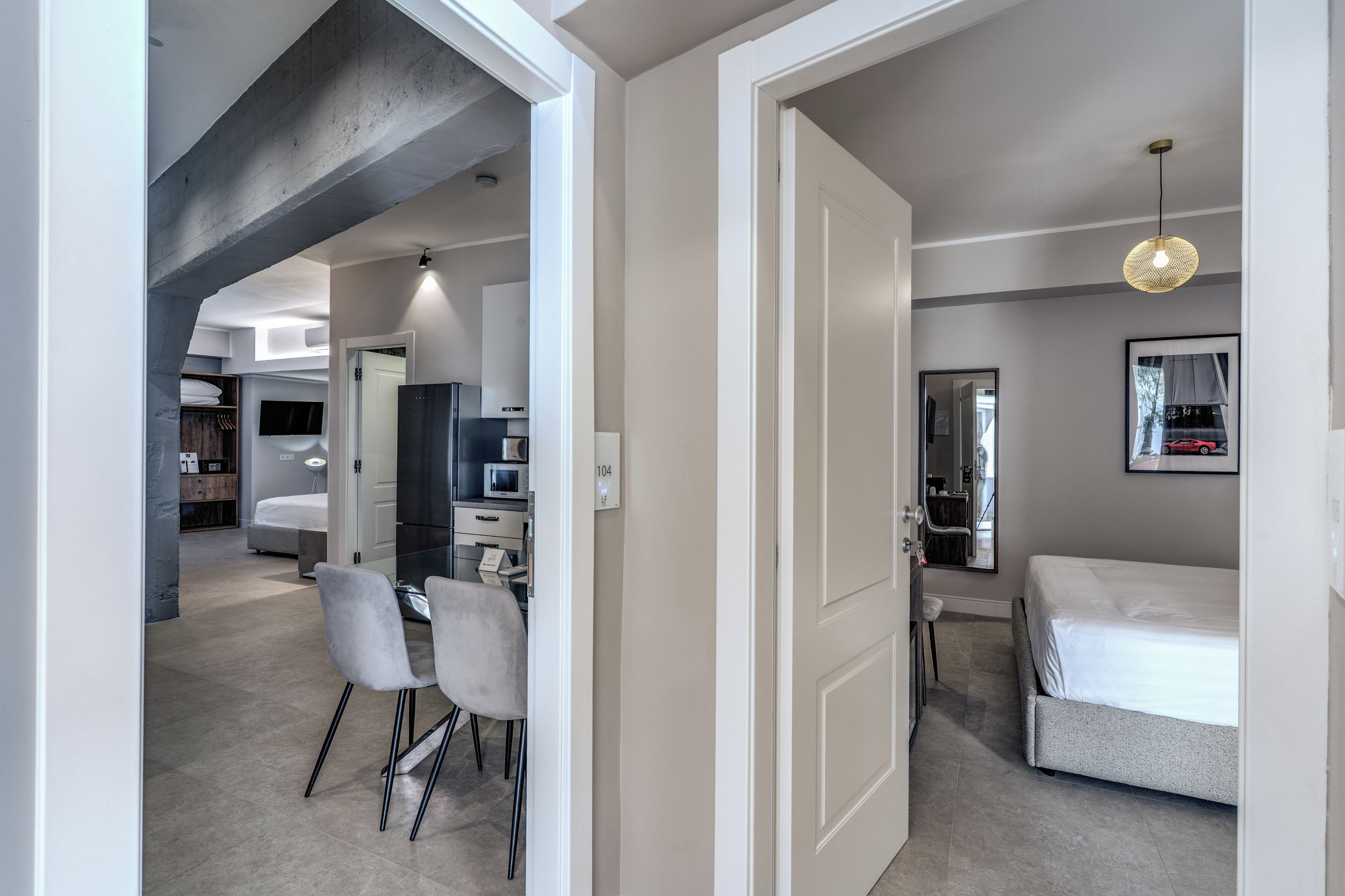 A modern bedroom with a bed, a desk, and chairs, a bathroom with a mirror, and a kitchen area with appliances.